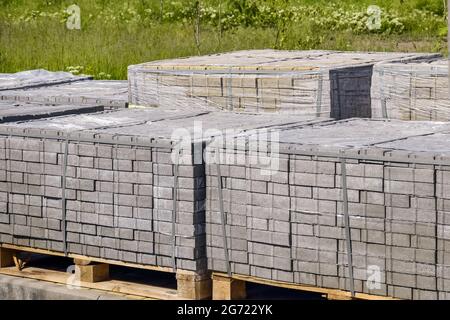 Paletten mit grauen Pflasterplatten auf dem Hintergrund von grünem Gras. Kunststoffverpackungen schützen die Fliese vor atmosphärischen Einflüssen. Stockfoto