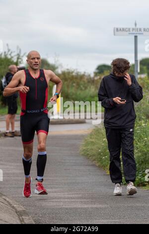 Shoeburyness, Essex, Großbritannien. Juli 2021. Aufgrund der COVID-19-Pandemie wurde der Triathlon 2020 verschoben, um wieder zurückzukehren, da die Beschränkungen für 2021 aufgehoben werden. Die Wettkämpfer, die im Shoeburyness-Gebiet östlich von Southend on Sea stattfinden, begannen den Wettbewerb mit einem 750-Meter-Schwimmen in der Themse-Mündung, bevor sie mit ihren Fahrrädern eine 20km-Fahrt durch Landstraßen und einen 5km-Lauf im Gunners Park an der Küste zurücknahmen Seine imposanten Verteidigungsstrukturen im Krieg. Das ehemalige Mod-Gebiet ist heute ein Naturschutzgebiet und beliebtes Wandergebiet. Läufer passiert einen jungen Mann, der sich auf das Telefon konzentriert Stockfoto