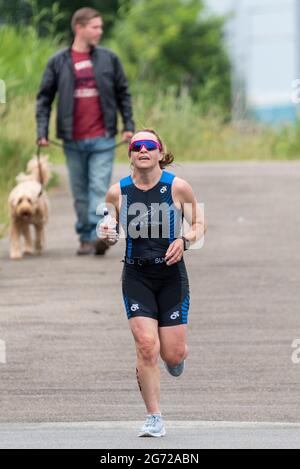 Shoeburyness, Essex, Großbritannien. Juli 2021. Aufgrund der COVID-19-Pandemie wurde der Triathlon 2020 verschoben, um wieder zurückzukehren, da die Beschränkungen für 2021 aufgehoben werden. Die Wettkämpfer, die im Shoeburyness-Gebiet östlich von Southend on Sea stattfinden, begannen den Wettbewerb mit einem 750-Meter-Schwimmen in der Themse-Mündung, bevor sie mit ihren Fahrrädern eine 20km-Fahrt durch Landstraßen und einen 5km-Lauf im Gunners Park an der Küste zurücknahmen Seine imposanten Verteidigungsstrukturen im Krieg. Das ehemalige Mod-Gebiet ist heute ein Naturschutzgebiet und beliebtes Wandergebiet. Läuferin, die an einem Hundespaziergänger vorbeikommt Stockfoto