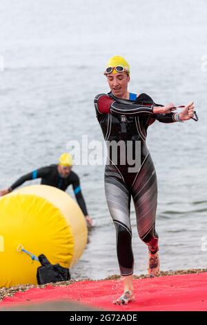 Shoeburyness, Essex, Großbritannien. Juli 2021. Aufgrund der COVID-19-Pandemie wurde der Triathlon 2020 verschoben, um wieder zurückzukehren, da die Beschränkungen für 2021 aufgehoben werden. Die Teilnehmer, die im Shoeburyness-Gebiet östlich von Southend on Sea stattfinden, begannen den Wettbewerb mit einem 750-Meter-Schwimmen in der Themse-Mündung, bevor sie ihre Fahrräder für eine 20-km-Fahrt durch Landstraßen und eine 5-km-Abfahrt im Gunners Park an der Küste abholten. Weibliche Sportlerin, die das Wasser verlässt Stockfoto