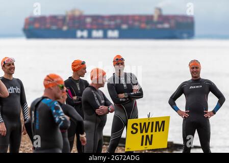 Shoeburyness, Essex, Großbritannien. Juli 2021. Aufgrund der COVID-19-Pandemie wurde der Triathlon 2020 verschoben, um wieder zurückzukehren, da die Beschränkungen für 2021 aufgehoben werden. Die Teilnehmer, die im Shoeburyness-Gebiet östlich von Southend on Sea stattfinden, begannen den Wettbewerb mit einem 750-Meter-Schwimmen in der Themse-Mündung, bevor sie ihre Fahrräder für eine 20-km-Fahrt durch Landstraßen und eine 5-km-Abfahrt im Gunners Park an der Küste abholten. Schwimmer am Start, während das Containerschiff HMM Danzig in die Nordsee fährt Stockfoto