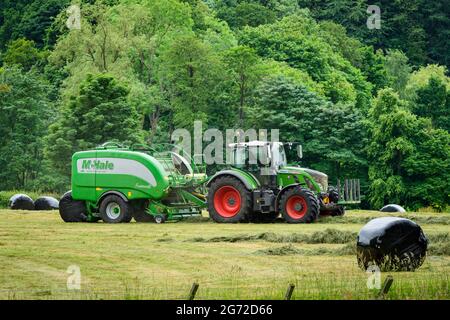 Heu- oder Silageherstellung (Landwirt im Traktor auf dem Bauernhof bei der Arbeit in ländlichen Feldern, Ballenpresse zu ziehen, trockenes Gras und runde Ballen einzusammeln) - Yorkshire England Großbritannien. Stockfoto