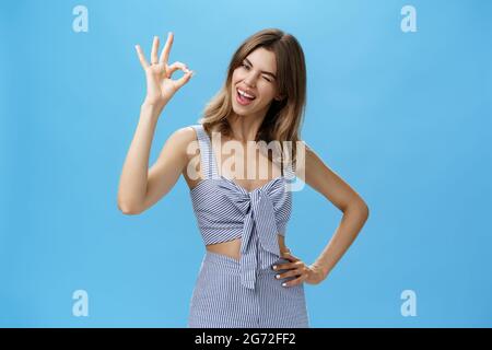 Nette charismatische Frau mit Lücke Zähne zeigt okay Geste kippenden Kopf freudig, die Hand auf der Taille und zwinkernd mit selbstbewussten Ausdruck Stockfoto