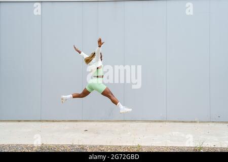 Seitenansicht einer afroamerikanischen Frau, die mit gespreizten Beinen und ausgestreckten Armen springt. Frau in Sportkleidung und weißer Jacke mit gelben Zöpfen springen Stockfoto