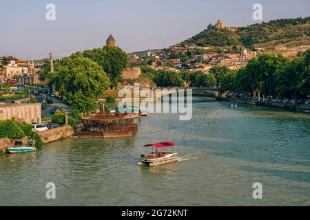 Tiflis, Georgien - 19. Juli 2019: Sommer Stadtbild bei Sonnenuntergang - Panoramablick mit Kura Fluss, bunte traditionelle Häuser, alte Brücke, touristische Boot Stockfoto