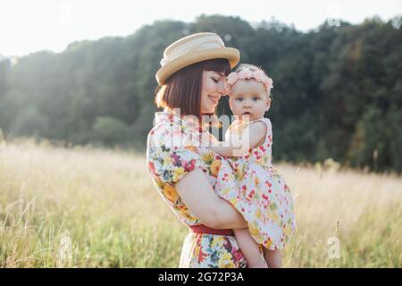 Lächelnde, fröhliche Mutter in Kleid und Hut, umarmt zärtlich die kleine Tochter, hat Spaß in der Natur, in den Strahlen des Sonnenuntergangs. Nettes Baby Mädchen lookis bei der Kamera Stockfoto