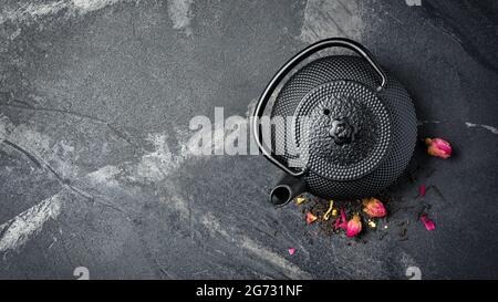 Draufsicht auf schwarzen asiatischen gusseisernen Wasserkocher und Tee mit Blütenblättern auf dunklem Marmorhintergrund mit Kopierfläche Stockfoto