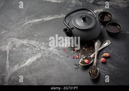 Schwarzer asiatischer gusseiserner Wasserkocher und Tee mit Blütenblättern in Vintage-Silberlöffeln auf dunklem Marmorhintergrund mit Kopierfläche Stockfoto