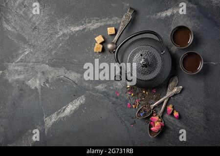 Draufsicht auf schwarzen asiatischen gusseisernen Wasserkocher und Tee mit Blütenblättern auf dunklem Marmorhintergrund mit Kopierfläche Stockfoto