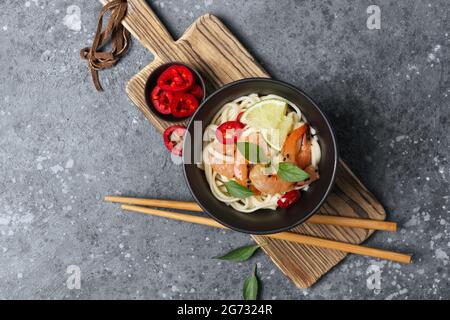 Draufsicht auf Schale mit japanischen udon-Nudeln, würzigen Garnelen, Limonen- und schwarzen Sesamkörnern auf Holzbrett und grauem Betonhintergrund Stockfoto