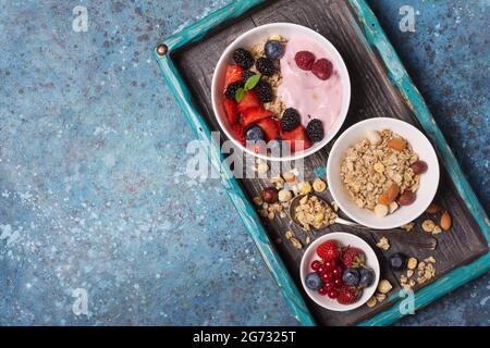 Draufsicht auf Schale mit Haferflocken, Himbeere, Erdbeere, Heidelbeere, rote Johannisbeere, Hausgemachter Joghurt aus Mandeln, Haselnüssen und Früchten auf türkisfarbenem Tablett und Blau Stockfoto