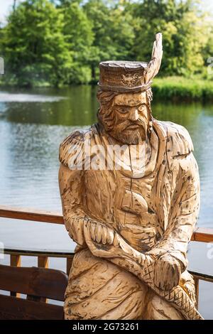 Zwierzyniec, Lubelskie, Polen - 02. Juli 2021: Holzskulptur eines traditionell gekleideten Mannes; schöner Teich im Hintergrund. Stockfoto
