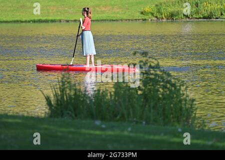 Eine junge Frau paddelt am 10. Juli 2021 im Olympiapark in München über den Olympiasee. Stand Up Paddling.SUP Stand Up Paddling (SUP), auch Stand Up Paddling oder Stand Up Paddle genannt, ist eine Wassersportart, bei der ein Athlet aufrecht auf einer Art Surfbrett steht und mit einem Paddel paddelt. Stockfoto