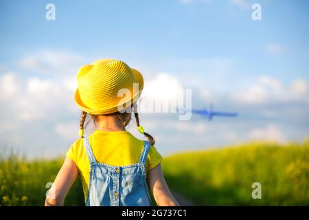 Mädchen in einem gelben Hut wirft ein Spielzeug Flugzeug auf das Feld, schaut auf die Spur. Sommerzeit, Kindheit, Träume und Nachlässigkeit. Flugtour von einer Reise Stockfoto