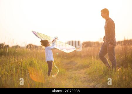 Papa hilft seiner Tochter, im Sommer bei Sonnenuntergang einen Drachen auf einem Feld zu fliegen. Familienunterhaltung im Freien, Vatertag, Kindertag. Ländliche Gebiete, zusatzbereich Stockfoto