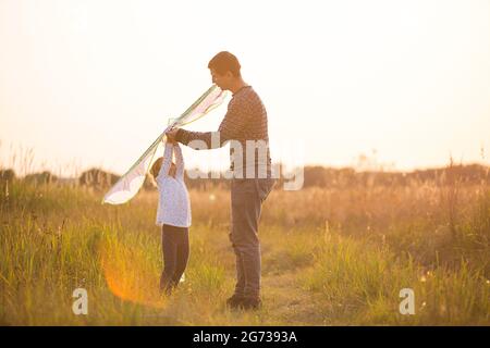 Papa hilft seiner Tochter, im Sommer bei Sonnenuntergang einen Drachen auf einem Feld zu fliegen. Familienunterhaltung im Freien, Vatertag, Kindertag. Ländliche Gebiete, zusatzbereich Stockfoto