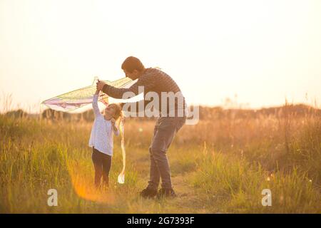 Papa hilft seiner Tochter, im Sommer bei Sonnenuntergang einen Drachen auf einem Feld zu fliegen. Familienunterhaltung im Freien, Vatertag, Kindertag. Ländliche Gebiete, zusatzbereich Stockfoto