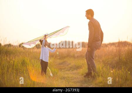 Papa hilft seiner Tochter, im Sommer bei Sonnenuntergang einen Drachen auf einem Feld zu fliegen. Familienunterhaltung im Freien, Vatertag, Kindertag. Ländliche Gebiete, zusatzbereich Stockfoto