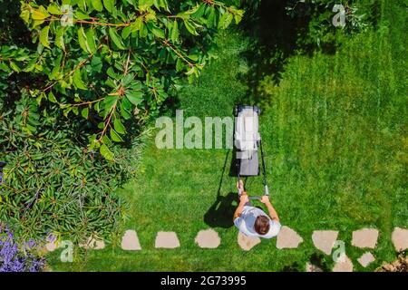 Kaukasischer Mann schiebt Rasenmäher für das Schneiden von grünem Gras im Garten mit Sonnenlicht und gutem Wetter in der Sommersaison. Luftaufnahme. Hausarbeit und Lebensmut Stockfoto