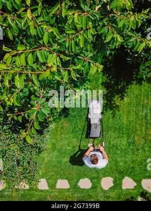 Kaukasischer Gärtner mäht Hinterhof-Gartengras mit Akku-Rasenmäher. Luftaufnahme. Garten- und Landschaftsbau. Stockfoto