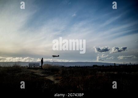 Silhouette eines Mannes, der neben einer Bank steht, mit einem kleinen Flugzeug, das tief über seinem Kopf fliegt, Berge am Horizont, blauer Himmel mit Wolken Stockfoto