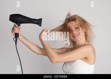 Schöne kaukasische Frau posiert mit nassem Haar. Frauen Haarbehandlung. Mädchen mit blonden Haaren mit Haartrockner. Frisur, Friseurkonzept. Stockfoto