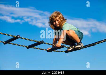 Netter Junge klettert die Leiter auf dem Spielplatz hoch. Kind klettert die Leiter gegen den blauen Himmel hinauf. Schöner lächelnder netter Junge auf einem Spielplatz. Kinder Stockfoto