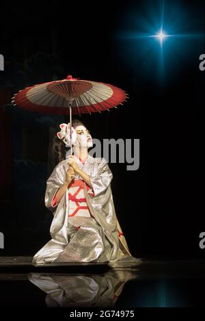 Geisha in traditionellem Kimono mit Regenschirm, der in der Dunkelheit auf den Knien sitzt, vom Stern aufgehellt und nach oben schaut. Traditionelle japanische Darbietung. Kin Stockfoto