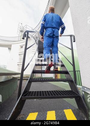 Manipulator im chemischen Betrieb trägt einen blauen Overall und geht eine Wendeltreppe aus Stahl in das obere Stockwerk hinauf. Stockfoto