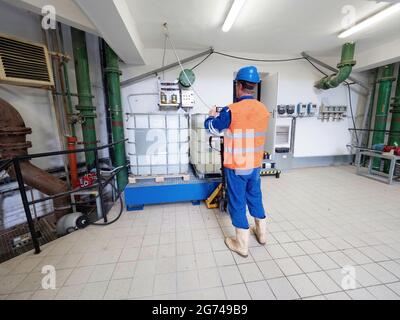 Manipulator im chemischen Betrieb mit reflektierender Weste, blauem Helm und gummierten Arbeitsstiefeln. Ein Mann handhabt einen Palettenwagen und ein chemisches Material Tan Stockfoto