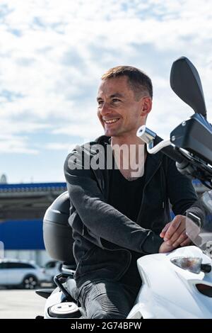 Attraktiver junger, stylischer lächelnder Mann, der auf einem Motorrad sitzt. Porträt eines hübschen Bikers, der in einer schwarzen Lederjacke auf einem Fahrrad posiert. Lifestyle-Phot Stockfoto