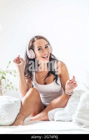 Das Mädchen sitzt in Hemd und Shorts auf dem Bett, umgeben von weißer Bettwäsche, hört die Musik über Kopfhörer und tanzt. Stockfoto