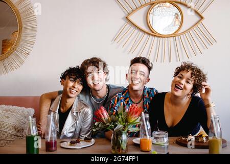 Kreis glücklicher Freunde, die in einem Restaurant zusammen sitzen. Vier junge, schwule Menschen lächeln beim Mittagessen fröhlich vor der Kamera. Freunde verbinden und s Stockfoto