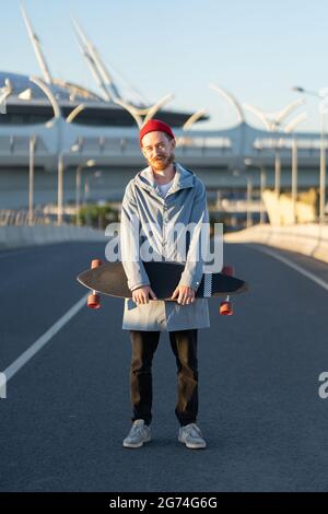 Junger bärtiger Hipster in rotem Hut hält Longboard in der City Street. Trendiges urbanes Lifestyle-Konzept Stockfoto