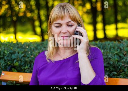 Unschärfe kaukasisch blonde Frau reden, sprechen am Telefon draußen, im Freien. 45er Jahre alte Frau in lila Bluse im Park. Erwachsene Frauen, die ein Mobiltelefon verwenden Stockfoto
