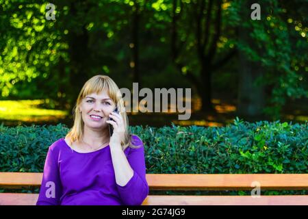 Unschärfe kaukasisch blonde lächelnde Frau sprechen, sprechen auf dem Telefon draußen, draußen. 40-jährige Frau in lila Bluse im Park auf der Bank. Erwachsene fe Stockfoto