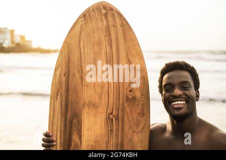 Black Surfer Mann hält vintage Surfbrett am Strand bei Sonnenuntergang - Fokus auf Gesicht Stockfoto