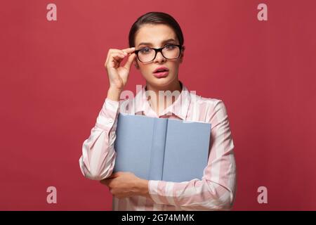 Porträt einer schockierten jungen Frau in einer Brille mit einem Buch mit leerem Einband auf rotem Hintergrund. Erstellen Sie ein Modell für den Kopierbereich. Menschen aufrichtige Emotionen Lifestyle-Konzept Stockfoto