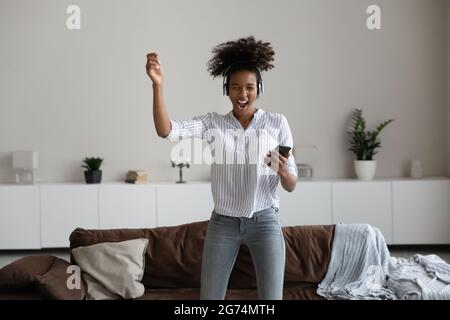 Glückliche tausendjährige afroamerikanische Frau in Kopfhörern tanzen, halten Smartphone Stockfoto