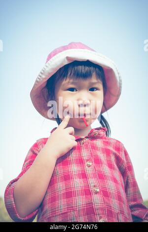 Nettes kleines asiatisches Mädchen, das im Sommer einen Lollipop auf Naturhintergrund isst. Kind trägt einen rosa Hut und schaut auf die Kamera. Prozessübergreifend. Vintage-Stil Stockfoto