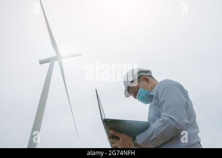 Ingenieure Windmühlen tragen Gesichtsmaske und arbeiten auf Laptop mit Die Windenergieanlage im Hintergrund Stockfoto