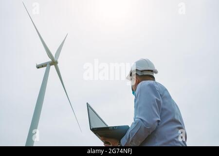 Ingenieure Windmühlen tragen Gesichtsmaske und arbeiten auf Laptop mit Die Windenergieanlage im Hintergrund Stockfoto