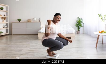 Aufgeregt kurvige schwarze Frau, die zu Hause auf einer Waage sitzt und YES Geste macht, glücklich über das Ergebnis ihrer Schlankheitskur Stockfoto