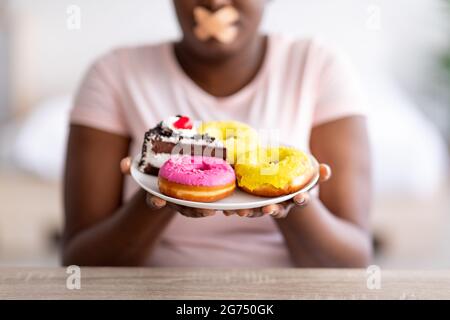 Nahaufnahme der kurvigen schwarzen Dame, die einen Teller mit Süßigkeiten hält, mit einer klebenden Bandage auf ihrem Mund, um die Gewichtsabnahme zu halten Stockfoto