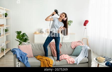 Indisches Teenager-Mädchen, das Kopfhörer trägt, den Fön als Mikrofon nutzt, zu Hause Gesang singt und auf dem Sofa tanzt Stockfoto