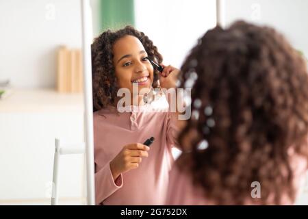 Make-up zu Hause. Über die Schulter des schwarzen Teenagers, das schwarze Mascara auftradet, den Spiegel anschaut und lächelt Stockfoto