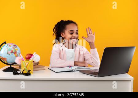 Portrait eines lächelnden schwarzen Mädchens, das am Tisch sitzt und mit der Hand winkt Stockfoto