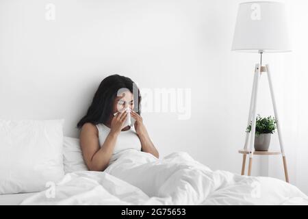 Krankheitstag zu Hause. Kranke afroamerikanische Frau hat laufende Nase, Husten und Erkältung, Nase blasen, im Bett sitzen, Platz kopieren Stockfoto