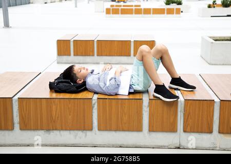 Müde Teenager Schuljunge schläft nach der Klasse auf dem Schulhof des Campus auf einer Holzbank, Schultasche unter seinem Kopf. Stockfoto