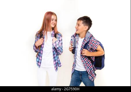 Junge und Mädchen Teenager 11 Jahre alt Schuljunge und Schülerin, Blick auf einander auf weißem Hintergrund mit Rucksäcken und lächelnd. Gekleidet in karierten Sh Stockfoto